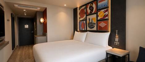 A hotel room with a large bed, a bedside table, a chair, a wall-mounted air conditioner, a door, and a wall with framed artwork.