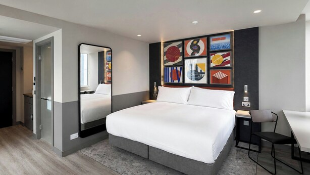 Habitación de hotel moderna con una cama grande, un escritorio y una silla. Se aprecia una pared con cuadros enmarcados y un espejo.