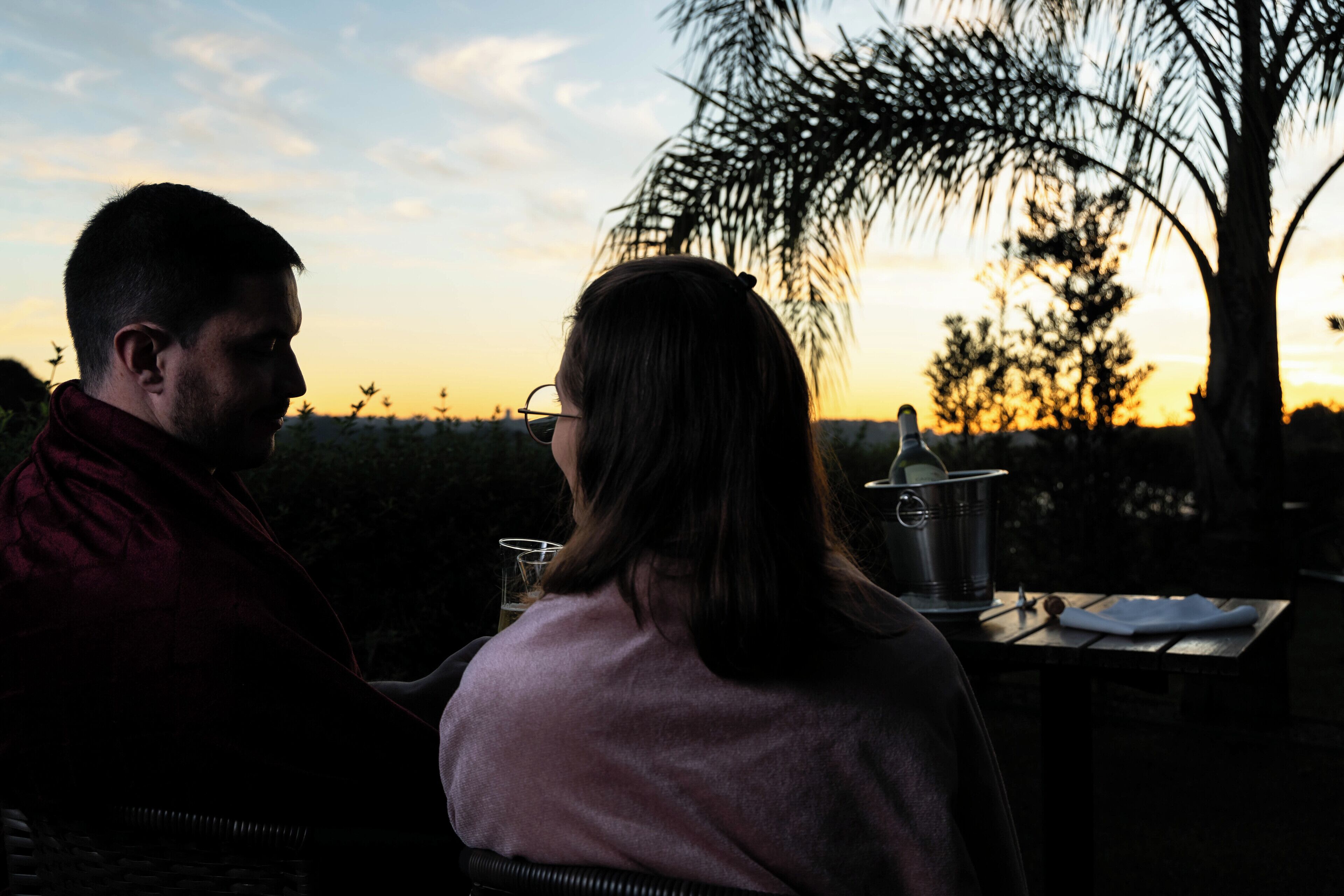 Un couple est assis à une table en plein air, avec un coucher de soleil en arrière-plan.
