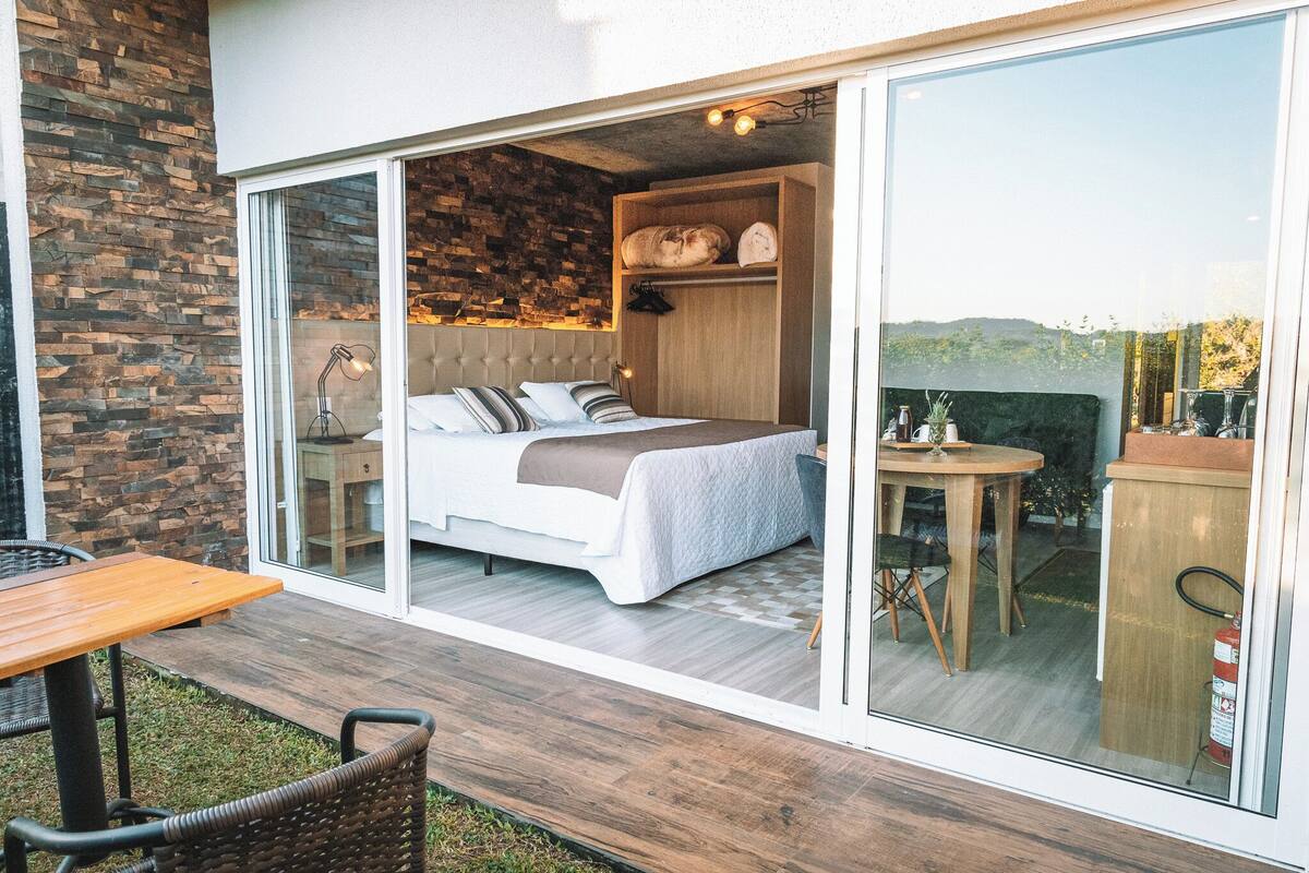 Ein Schlafzimmer mit Bett, Nachttisch und Blick durch eine Glastür auf einen Außenbereich mit Tisch und Stühlen.