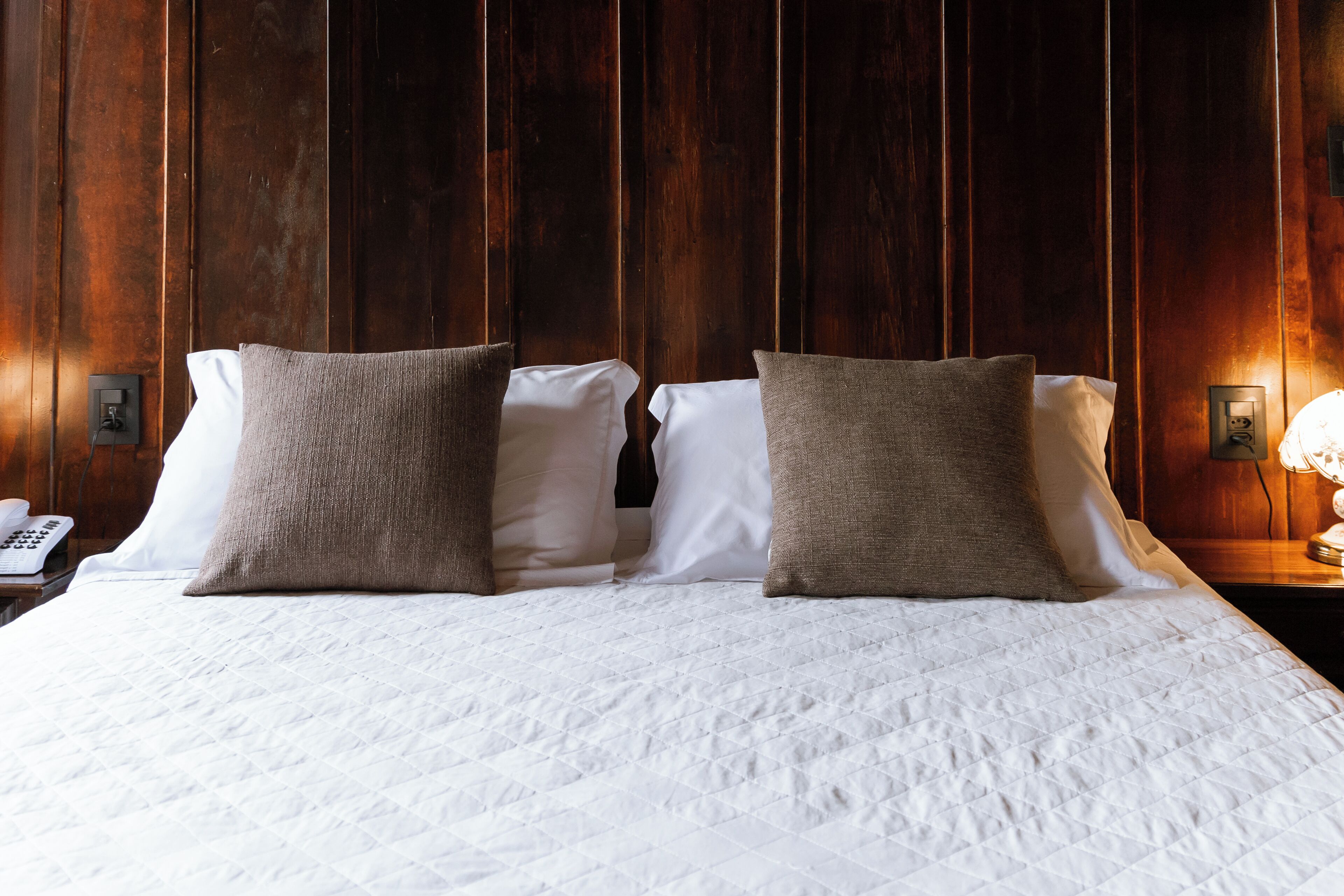 A neatly made bed with white bedding and two brown pillows, a bedside telephone, and a wall-mounted light fixture.