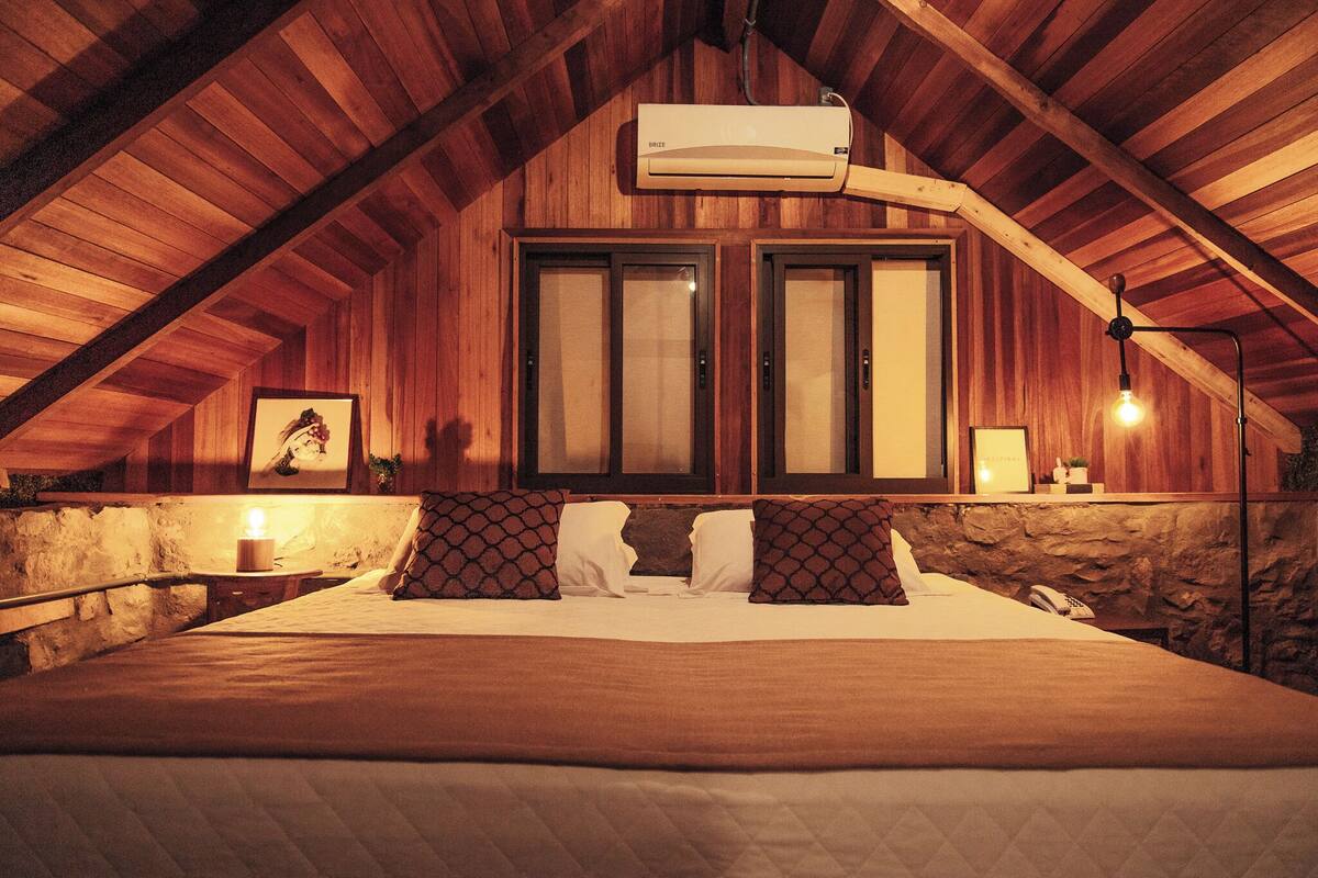 Ein gemütliches Schlafzimmer unter dem Dach mit Holzbalkendecke, einem großen Bett mit gemusterten Kissen, einer Steinwand und einer Wandmontage-Klimaanlage.