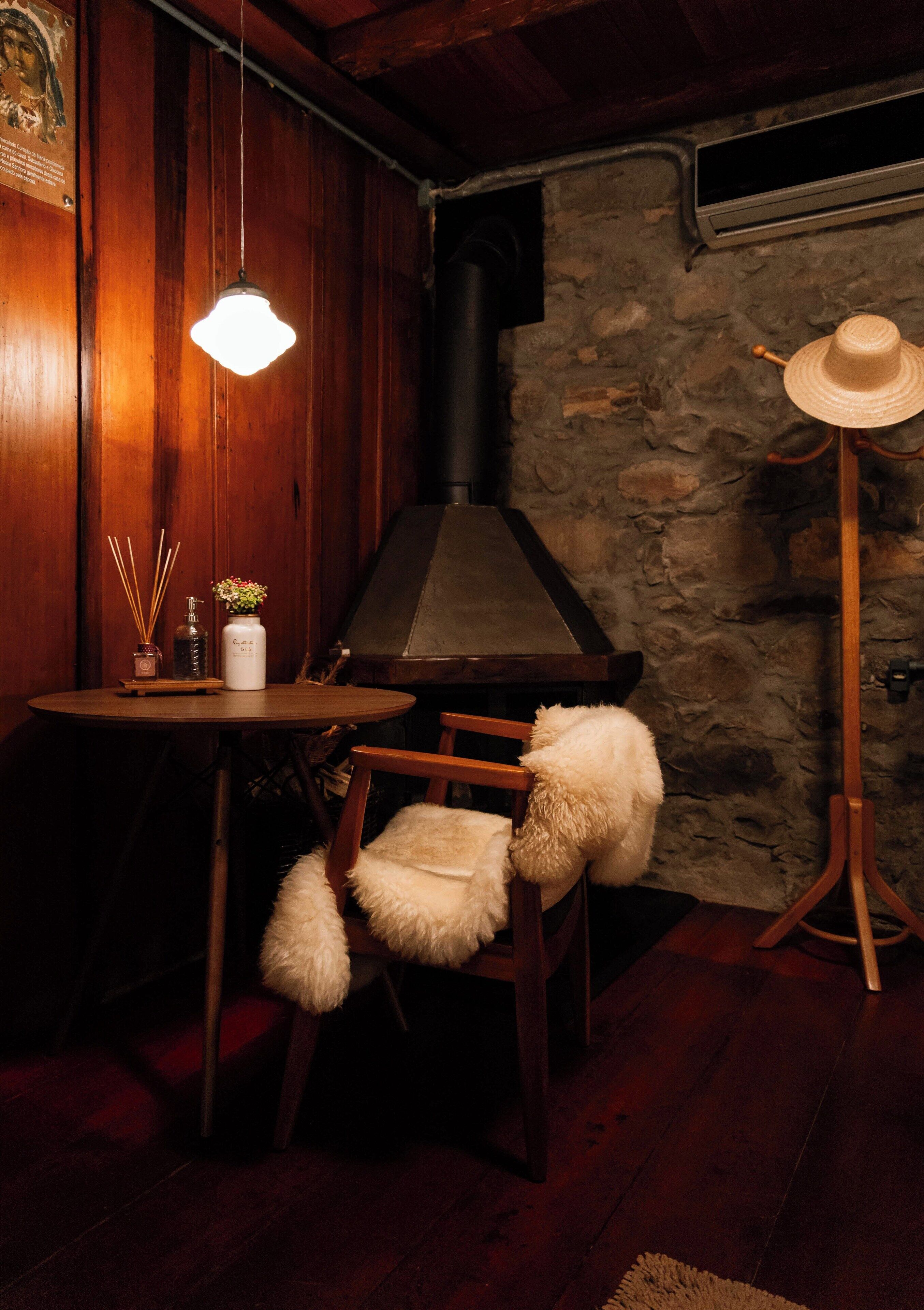 Un cuarto acogedor con pared de piedra, una mesa de madera con un florero y difusor de cañas, un sillón con cojín mullido y una lámpara de pie.