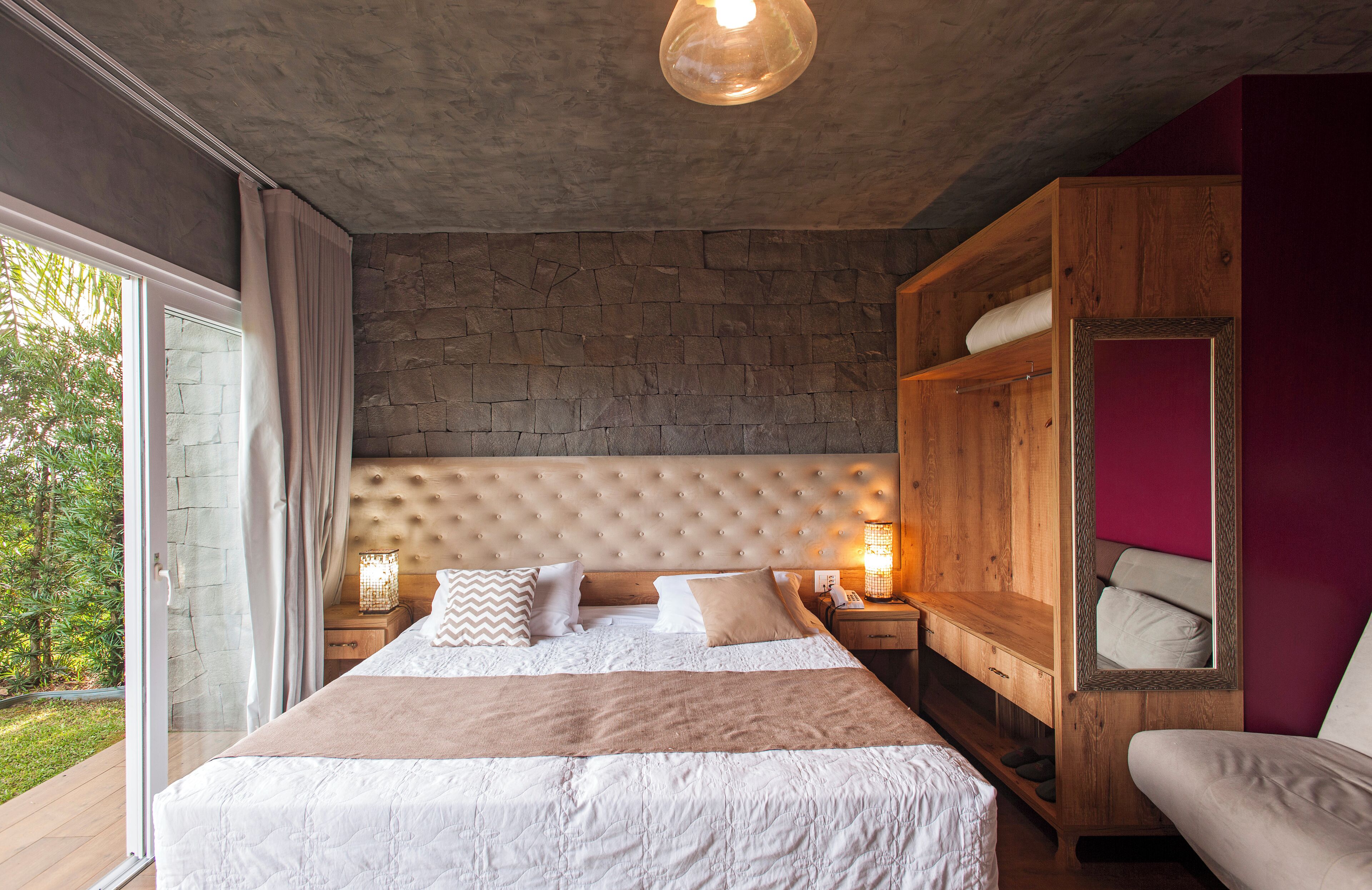 A bedroom with a large bed, a wooden wardrobe, a nightstand with a lamp, and a view of greenery outside.