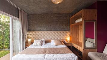 A bedroom with a large bed, a wooden wardrobe, a nightstand with a lamp, and a view of greenery outside.