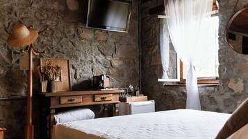 A rustic room with a stone wall, wooden furniture, a bed with white bedding, a television mounted above a dresser, and a window with sheer curtains.
