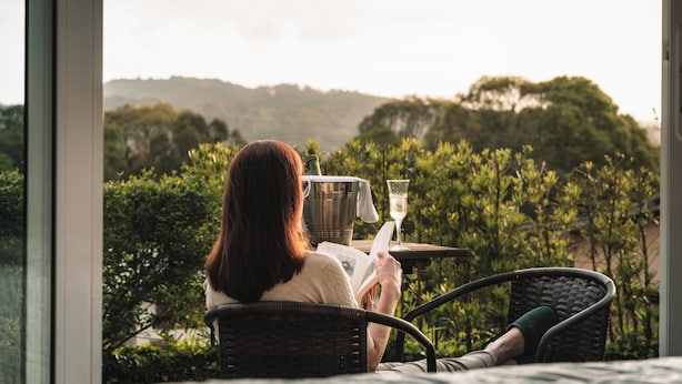 Une personne assise sur une chaise, lisant un livre, avec un verre de vin sur une table, et devant elle, un jardin luxuriant et des collines.