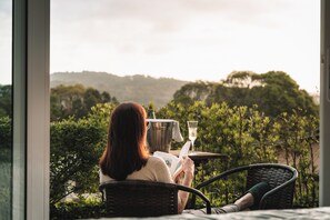 Uma pessoa sentada em uma cadeira lendo um livro, com uma taça de vinho sobre a mesa, apreciando um jardim exuberante e colinas.