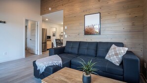 Una sala de estar moderna con un sofá seccional azul oscuro, una mesa de centro de madera con una planta, y una pared revestida de madera con un cuadro enmarcado.