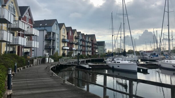 Ein Yachthafen mit vertäuten Booten, eine Holzpromenade, die zum Wasser führt, und farbenfrohe Wohnhäuser entlang der Küste.