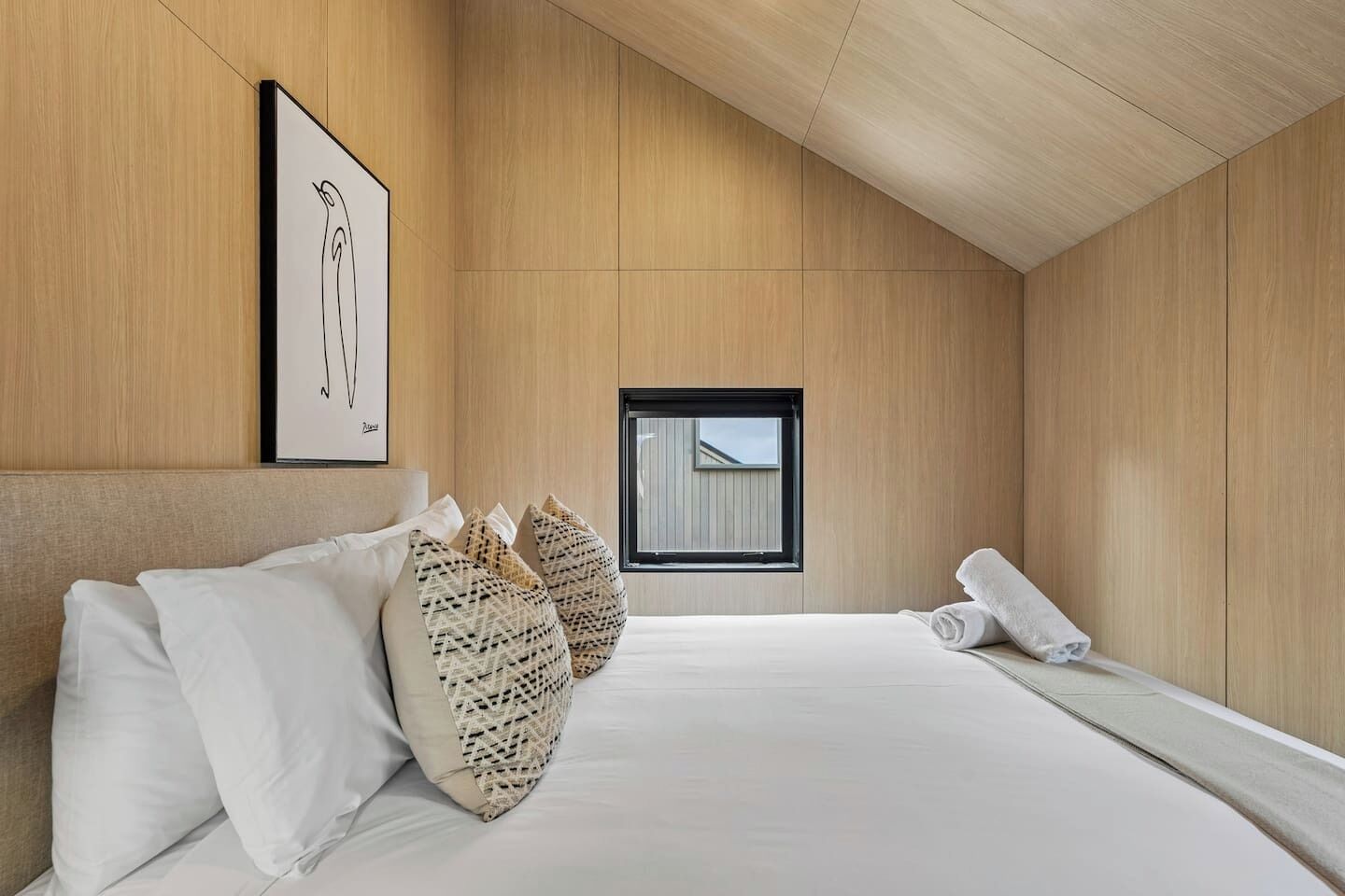 A modern bedroom with a wooden ceiling, a large bed with white linens, patterned pillows, a framed artwork on the wall, and a window with a view of a building.