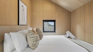 A modern bedroom with a wooden ceiling, a large bed with white linens, patterned pillows, a framed artwork on the wall, and a window with a view of a building.