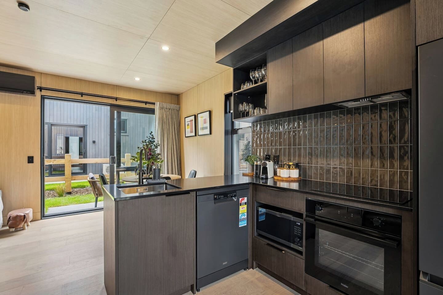 A modern kitchen with a built-in oven, a microwave, and a coffee machine. There is a dining area with a table and chairs visible through a glass door.
