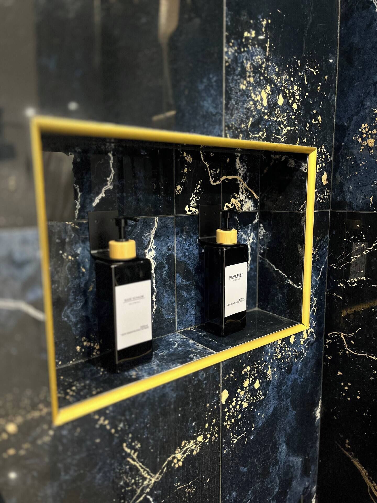 Ein Badezimmer mit schwarzen Marmorfliesen und einem goldfarbenen Spiegel. Unter dem Spiegel befindet sich ein Regal mit zwei Seifenspendern.