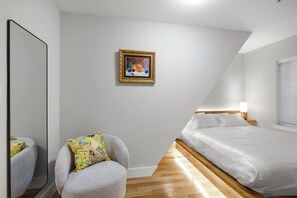 Un dormitorio con cama, una silla, mesita de noche con lámpara, un espejo grande y un cuadro en la pared.