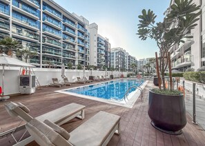 En poolanläggning på taket med solstolar, en stor blomkruka och moderna bostadshus i bakgrunden.