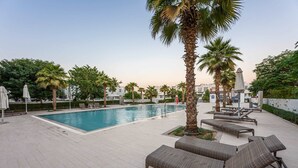 A poolside area with lounge chairs, palm trees, and a clear blue pool. - Stayshort - Modern Villa in Avencia Damac Hills 2 With Waterpark Access (Dubai)