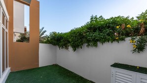 A small enclosed patio area with artificial green turf, a white wall, and a section of climbing plants with yellow flowers. - Stayshort - Modern Villa in Avencia Damac Hills 2 With Waterpark Access (Dubai)
