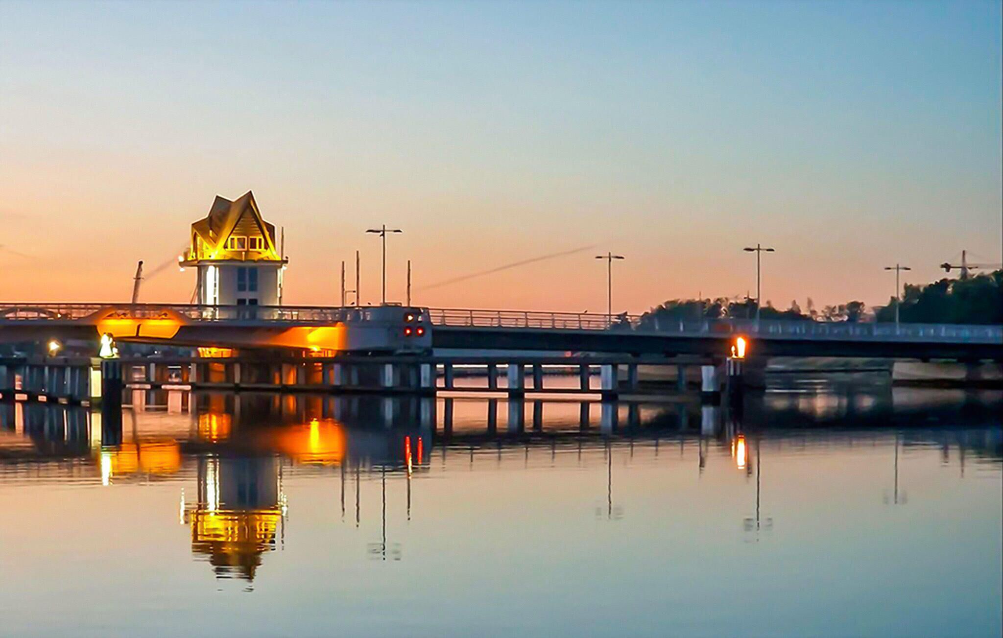 Une jetée bordant le rivage, surmontée d’un phare à son extrémité, et dont la surface d’eau calme reflète la structure.