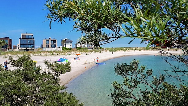 Ein Strand mit klarem türkisblauem Wasser, sandigem Ufer und modernen Gebäuden im Hintergrund.