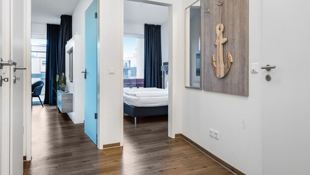 Habitación de hotel con cama, escritorio y silla. Hay una ventana con cortinas y una puerta que da a otra habitación.