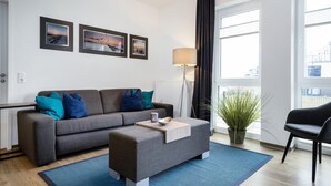 Una sala de estar moderna con un sofá gris, un reposapiés a juego, una lámpara de pie y un ventanal con cortinas.
