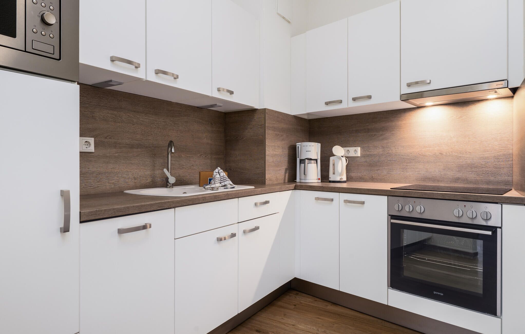 Eine moderne Küche mit weißen Schränken, einer Holzrückwand, einem Einbauofen, einer Kaffeemaschine und einem Spülbecken.