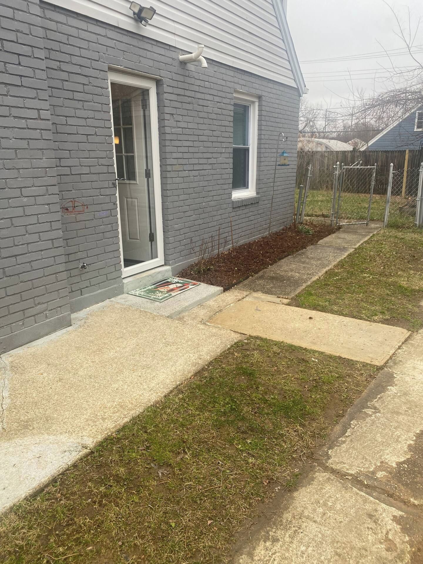 A residential house with a grey brick exterior, a front door, a small window, and a concrete pathway leading to the door.