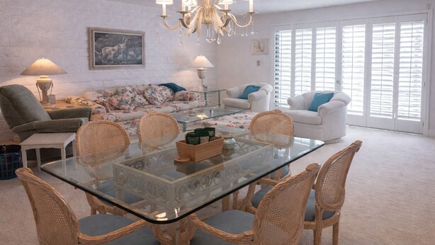 A spacious living room with a glass-top dining table surrounded by wicker chairs, a white sofa, and a white armchair. A chandelier hangs from the ceiling, and there is a large window with blinds. - 2-bedroom condo in fantastic Cathedral City with AC (Cathedral City)