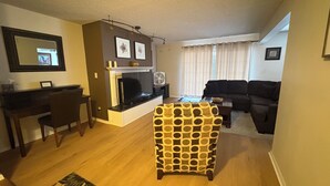 Una sala de estar con chimenea, un sofá, un sillón estampado, una zona de comedor con un escritorio y una silla, y una pequeña alfombra.