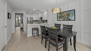 Une cuisine moderne avec des meubles blancs, un îlot central et un coin repas comprenant une table et des chaises aux couleurs sombres.