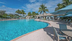 Une piscine d’un bleu éclatant, bordée de chaises longues et d’ombrelles, entourée de palmiers et surplombée par un ciel dégagé.