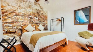 Ein Schlafzimmer mit Backsteinwand, einem Bett mit Kissen, einer Pendelleuchte, einem Sessel und einem Fenster mit Vorhang.