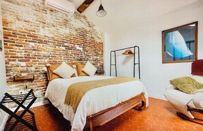 Quarto com parede de tijolos, cama com almofadas, luminária pendente, cadeira e janela com cortina.