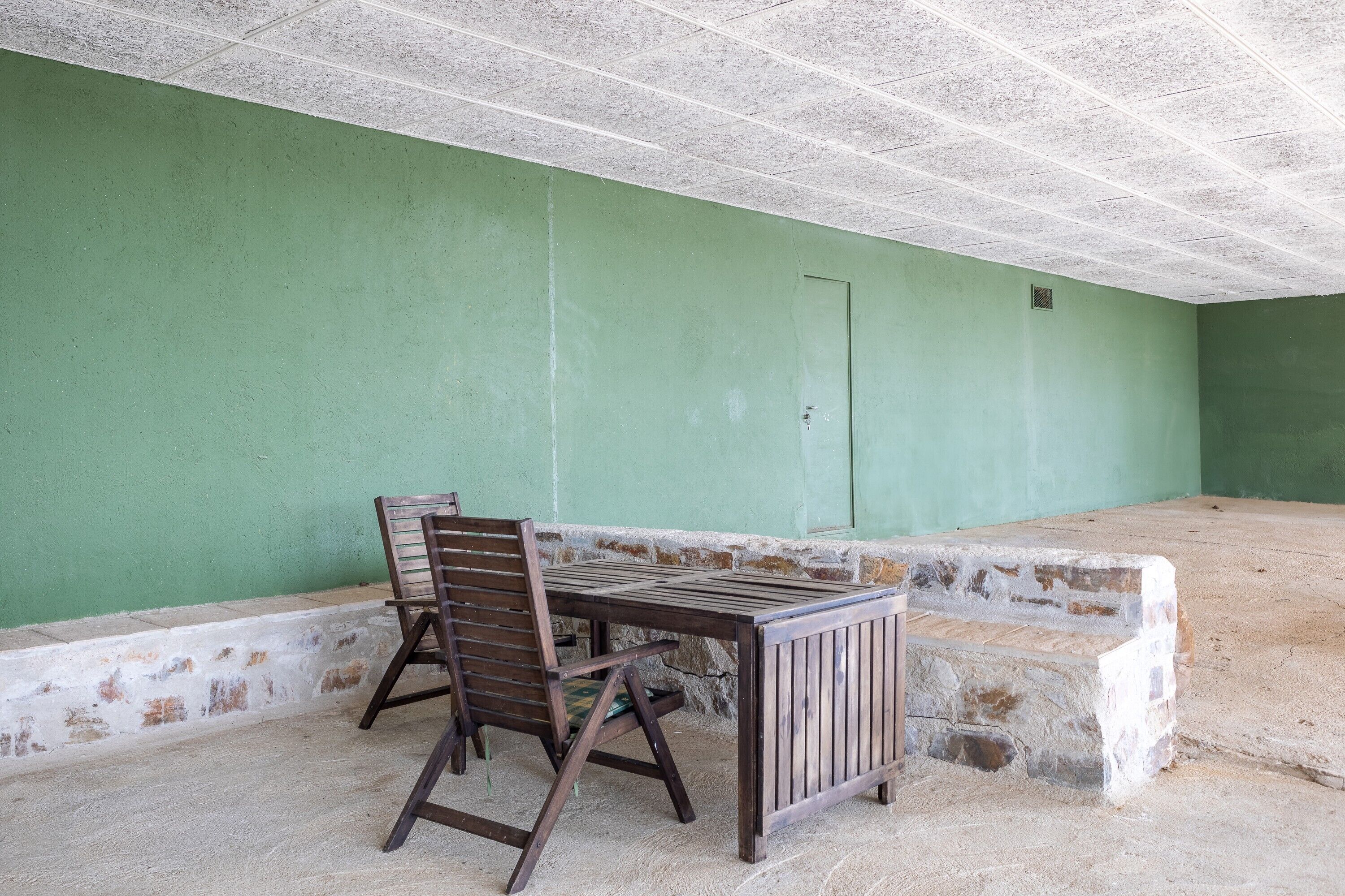 Una habitación con pared verde, una mesa de madera con una silla y piso de concreto.