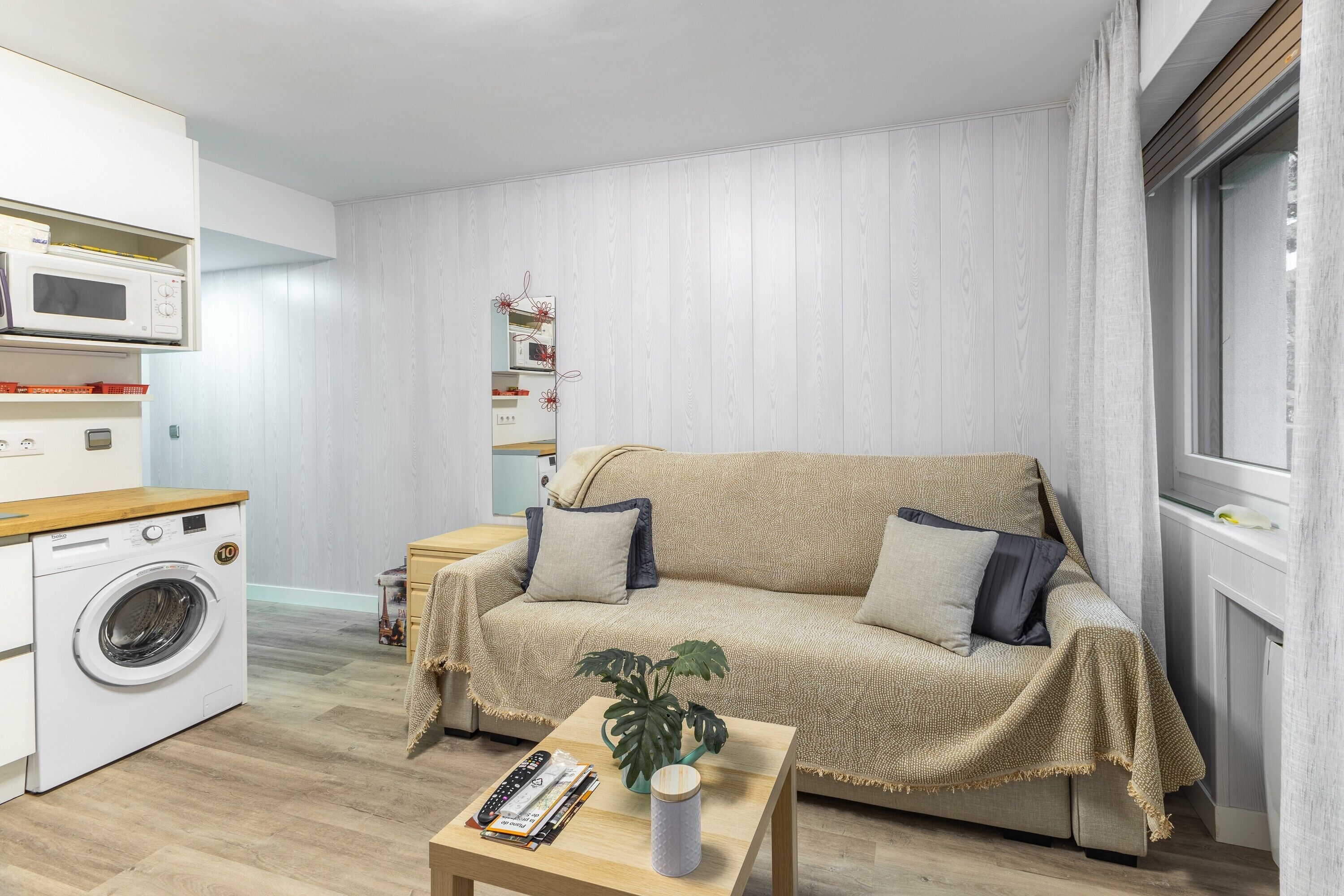Un espacio habitable compacto con lavadora, sofá, una mesita con planta y ventana con cortinas.