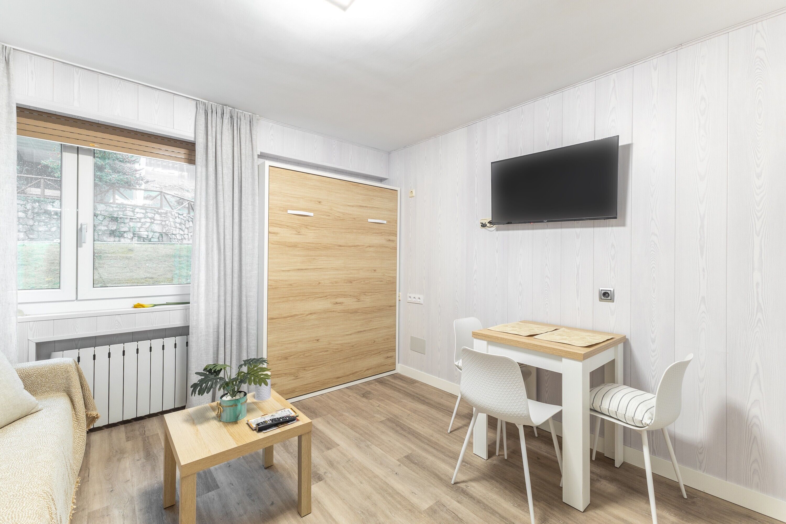 Una habitación con piso de madera, un sofá blanco, una pequeña mesa de madera con una planta, un televisor montado en la pared y una ventana con cortinas.