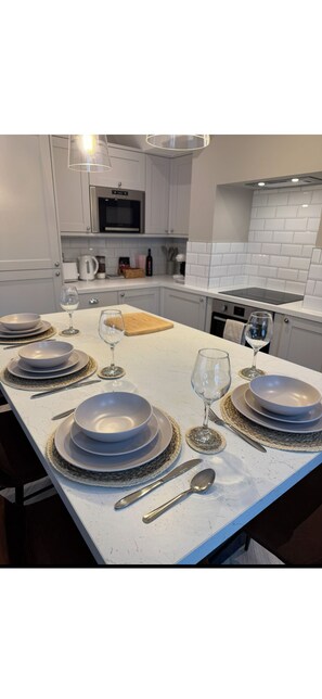 Una cocina moderna con armarios blancos, una isla central con tabla de cortar de madera, y una zona de comedor con platos azules y cubiertos.