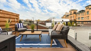 A rooftop terrace with outdoor furniture, a blue mat, and a view of surrounding buildings. - Rooftop Deck | 3 King Suites | Soaker Tub (Pittsburgh)