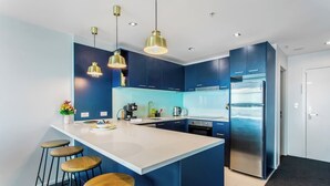 Eine moderne Küche mit blauen Schränken, einer weißen Kochinsel und Edelstahlgeräten. Drei Barhocker und Pendelleuchten befinden sich über der Kochinsel.