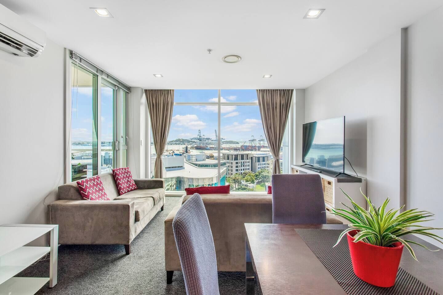 A modern hotel room with a large window, a sofa, a chair, a dining table, and a TV.