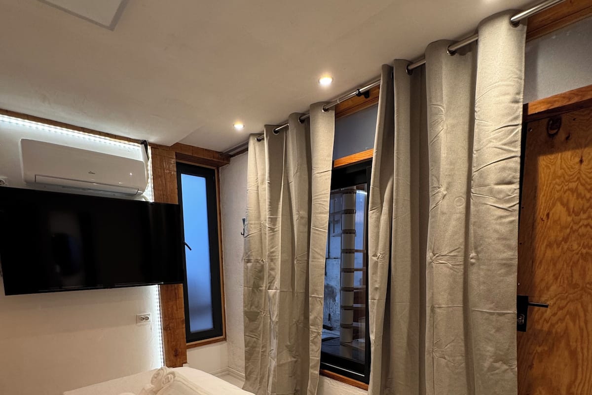 A bedroom with a bed, a flat-screen TV mounted on the wall, a window with curtains, and a wooden door.