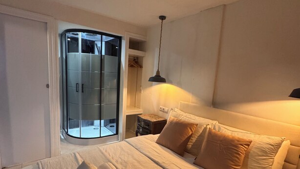 A neatly made bed with a white duvet and pillows, a bedside table with a lamp, and a sliding glass door leading to a bathroom. - WhatsApp  (Barcelona)