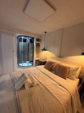 Een netjes opgemaakt bed met een witte deken en kussens, een nachtkastje met een lamp, en een schuifdeur naar een badkamer.