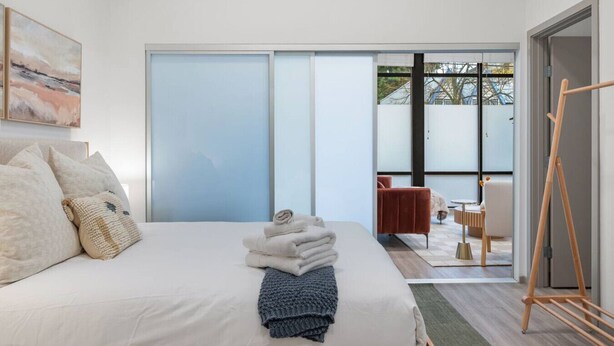 A modern bedroom with a large bed, a view of the outside through a sliding door, and a wooden easel in the corner. - Roosevelt Suite| Walk to Light Rail + Rooftop View (Seattle)