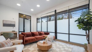 A modern living room with a brown sofa, white armchairs, a wooden coffee table, and large windows with blinds. - Roosevelt Suite| Walk to Light Rail + Rooftop View (Seattle)