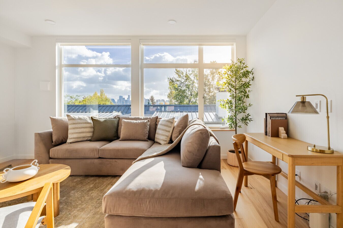 A modern living room with a sectional sofa, a wooden desk, and a large window offering a city view.