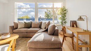A modern living room with a sectional sofa, a wooden desk, and a large window offering a city view. - NEW Queen Anne View Retreat | Sleeps 8 w/ A/C (Seattle)