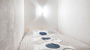 Una habitación con una cama cubierta por una manta blanca y azul estampada, una pared blanca y una lámpara en la pared.