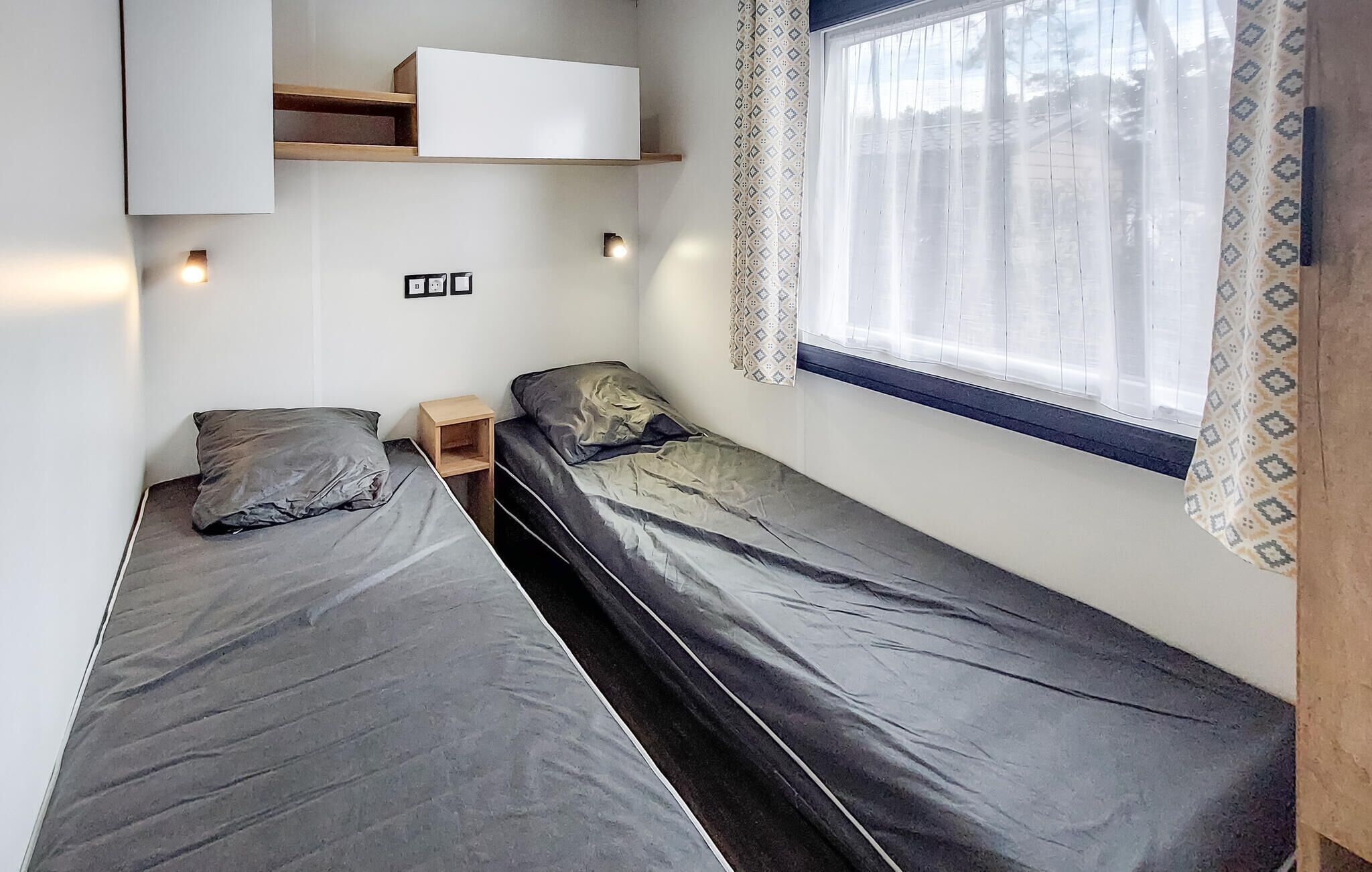 A small, single-bed room with a wooden shelf above the bed and a window with patterned curtains.